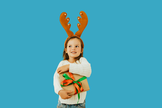 Adorable little girl in reindeer antlers holding a festive gift with ribbon, wearing white sweater and jeans. Child smiling against blue background, holiday celebration concept.