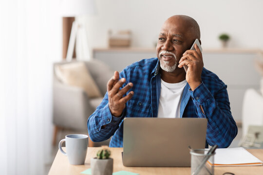 A senior African American businessman is talking on his mobile phone while seated at a laptop in a bright office.