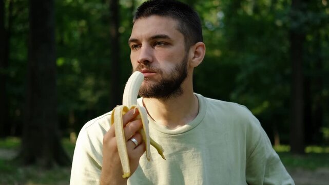Young man in light green t shirt eating banana outdoors while sitting in green park surrounded by trees and sunlight. 4k horizontal footage