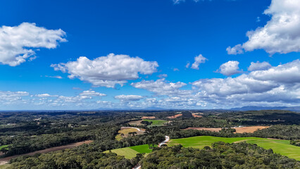 A panoramic rural landscape with hills covered in forests and green and brown fields. The sky is blue with white clouds, and sunlight illuminates the scene.