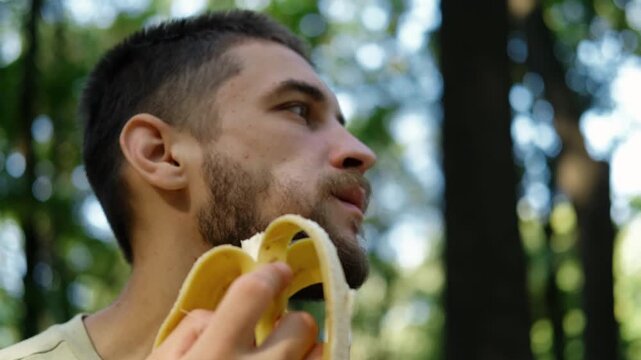 Young man in light green t shirt eating banana outdoors while sitting in green park surrounded by trees and sunlight. 4k horizontal footage