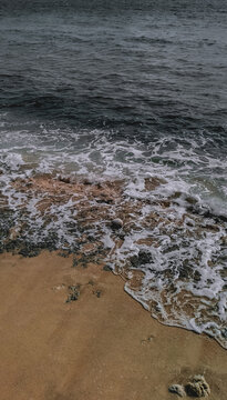 High-angle view of moody dark waves crashing onto the unique orange-brown sand and mossy rocks of Dato Beach, Majene, West Sulawesi. A perfect texture and pattern for coastal and abstract themes.
