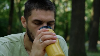 Young man in light green t shirt sitting outdoors among trees, drinking hot coffee or tea from yellow thermocup. 4k horizontal footage