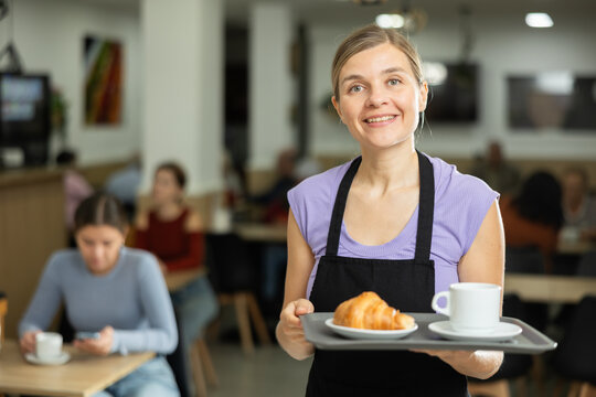 Polite waitress holding tray with coffee and croissant for clients in cozy cafeteria