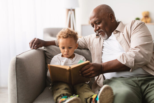 An African American grandfather sits on a sofa with his little grandson, sharing a book. The two focus intently on reading together, creating a warm family moment during the weekend. - Powered by Adobe