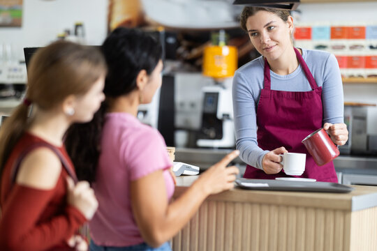 Polite young barista pouring coffee into cup while clients waiting at counter in cafeteria - Powered by Adobe