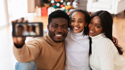 Obraz premium A happy black family enjoys quality time together, celebrating Christmas at home. They take a cheerful selfie with their excited daughter hugging her parents, all smiling and looking at the camera.