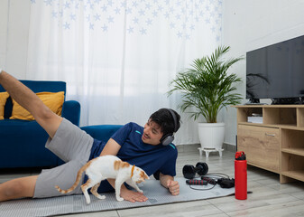 Man training at home on a mat, performing a lateral leg raise while his adorable pet plays beside him. Concept of home workout, well being, and active lifestyle