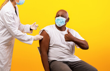 A senior black man receives a coronavirus vaccine shot from a medical doctor. He is wearing a face mask and shows a thumbs up, recommending vaccination against COVID-19.
