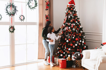 Loving black family members joyfully decorate a festive Christmas tree in their warm living room. A...
