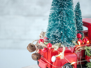 Old red country toy truck and twinkle fairy lights with holiday trees and festive Christmas decorations on white light backdrop. Vintage sleigh bells, greenery wreath, and red berries.