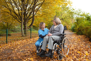 Elderly happy woman in wheelchair on a walk though park covers in fallen leaves with healthcare worker or nurse in uniform