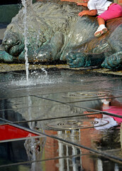 dziecko siedzące na kwamiennym lwie przy fontannie, odbicie dziecka w wodzie, a small child sitting on a stone lion, a child and water splashes in city fountain, summer children pastime   © kateej
