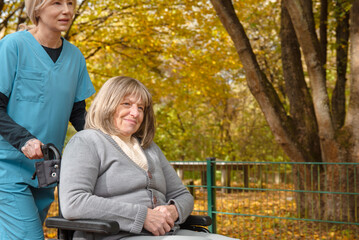 Fototapeta premium Elderly happy woman in wheelchair on a walk though park covers in fallen leaves with healthcare worker or nurse in uniform