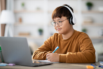 Cheerful korean schoolboy attends an online lesson from his home. He uses a laptop and takes notes in his notebook, enjoying the learning experience.