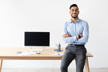 A smiling bearded business man leans on a table in his home office, confidently displaying a laptop...