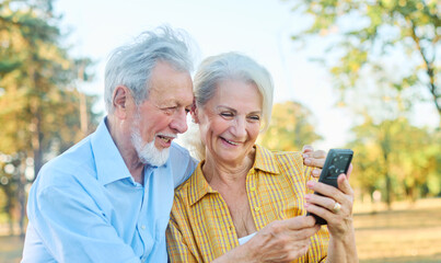 Happy active senior couple having fun using smart phones sitting on a bench outdoors