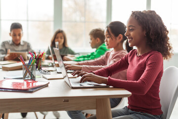 Black schoolgirl uses laptop while seated at a desk in a classroom surrounded by multiethnic classmates. The students focus on their screens, participating in online learning together.