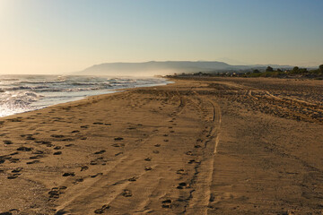Tranquil beach scene at sunset with gentle waves lapping against sandy shore, showcasing footprints and distant mountains in the background