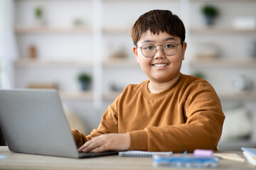 Chubby smart boy is focused on his homework at a table in a well-lit room. He types on his laptop while surrounded by school supplies, engaging in online learning.