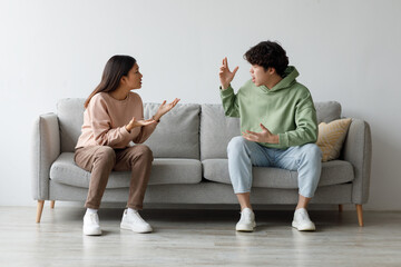 A young Asian couple is sitting on a grey sofa in their living room, visibly upset and yelling at each other. Emotions run high as they express their frustrations and disagreements.