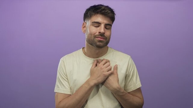 Young hispanic man in beige t shirt with light stubble places both hands on chest in studio with purple backdrop and calm expression; gratitude.