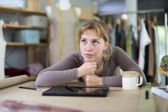 Thoughtful young female dressmaker sitting at table with tablet on it in sewing workshop