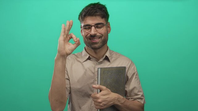 Smiling man wearing glasses holds book and makes ok gesture with hand in bright green studio; approval.