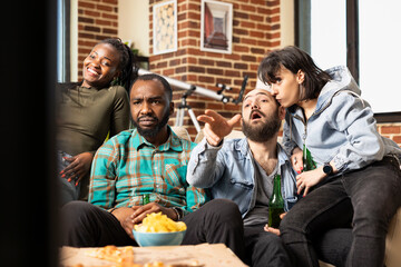 Group of friends sitting on couch, watching interesting movie on television, casual weekend at home. Multiethnic individuals spending time together in cozy apartment, having snacks and drinks nearby.