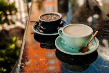 Black and white coffee cups on marble table. Morning cappuccino made by professional barista. Drawings on surface of coffee drinks. Hot chocolate with nuts in dark porcelain cup and teaspoon