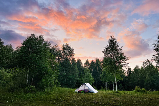 White tourist tent with camping gear in grassy clearing against backdrop of dense trees. Sky glows with vibrant hues of orange and pink as sun sets, casting serene ambiance over peaceful camping site.