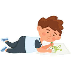 Young boy with brown hair wearing a blue and white shirt and dark pants lying on the floor drawing a green picture on white paper with a green crayon a happy 