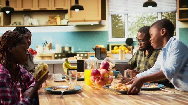 Black working dad on tablet ignoring son and family at breakfast table, absorbed disconnected man distracted by his remote work. Inattentive parent distant with his children and wife. Camera A.
