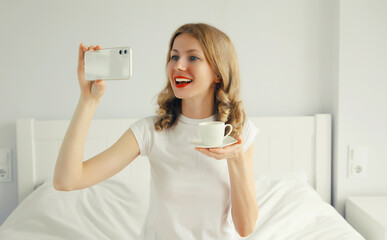 portrait of happy smiling young woman taking selfie with phone while lying on bed at home