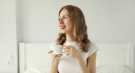 Happy smiling young woman enjoying with cup of hot coffee lying on bed at home