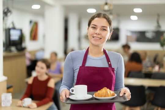 Young female waiter with tray of fresh coffee and croissant in cafe