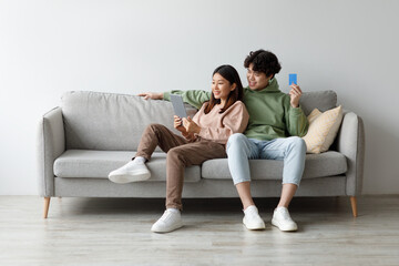 Cheerful Asian man and woman sit comfortably on a grey sofa at home, browsing online shops together. They are smiling, holding a credit card and a digital tablet, clearly enjoying the experience.