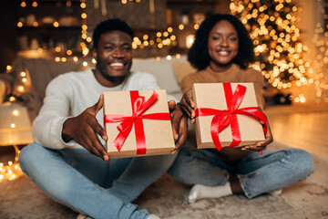 A joyful young black couple sits on the floor, holding Christmas presents towards the camera.