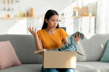 Fotobehang Muziek In a bright living room, a young asian woman sits on a couch, opening a box. She looks frustrated and unhappy after finding the wrong shirt inside the package she ordered online.  © Prostock-studio