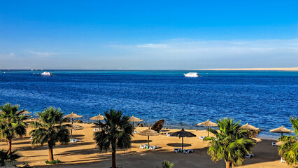 Obraz premium Scenic beach view with palm trees and umbrellas overlooking tranquil blue waters, boats in the distance, creating a serene coastal atmosphere for relaxation and leisure. Hurghada, Egypt, Red Sea
