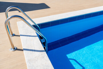 Closeup of pool ladder and tiles. sunlit blue water, stainless steel ladder, concrete deck, crisp geometric lines, inviting entry for swimmers, summer staycation