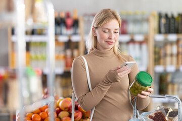 Girl scan QR code, take reading pickled olives label using phone camera in shop. Client inspect olive package product, view manufacturer information using mobile application