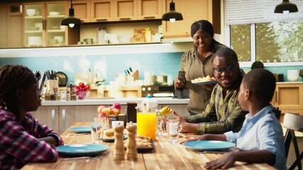 Happy black family enjoying breakfast together in kitchen with delicious food. Mother serves eggs, toast and orange juice while children smile, sharing joy of relaxed weekend morning. Camera A. - Powered by Adobe