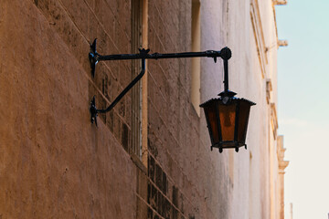A dark metal, antique wall lantern hangs from a bracket against a textured, sunlit stone building facade, casting a warm orange light.