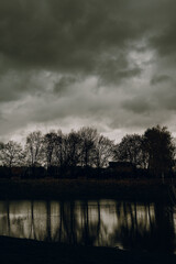 Dramatic dark autumn landscape with bare trees and cloudy sky reflecting in calm lake water. Moody nature background for fall season, overcast weather