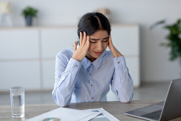 A young Indian woman sits at her workplace, visibly upset and holding her head. She appears to be...