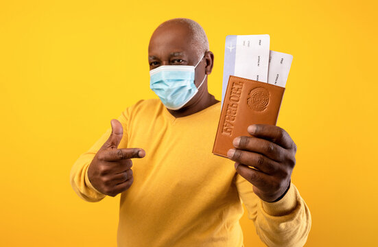 Elderly black man wearing a medical mask holds plane tickets and a passport while pointing confidently. This scene captures the excitement of travel during the coronavirus pandemic.