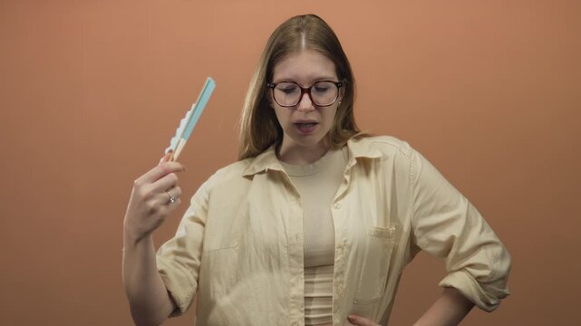 Woman wearing glasses with furrowed brow waves a turquoise folding fan in steady motion in studio; discomfort heat fatigue exhaustion.