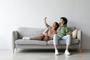 Young Asian couple sits comfortably on a couch, using a remote control to adjust the split air...