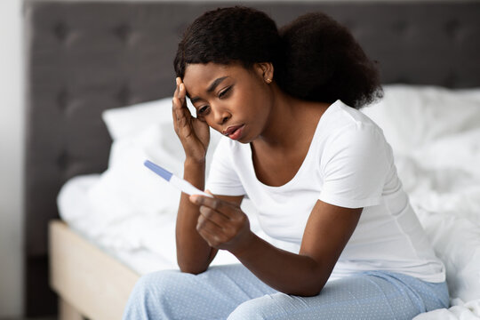 A single black woman in pajamas sits on her bed, holding a pregnancy test with a look of confusion on her face. She touches her head, reflecting on her unexpected situation at home.